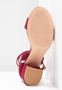 Sandale à talons hauts en daim rouge avec des brides entrecroisées, un bout pointu et une semelle en caoutchouc beige texturé présentant un motif ondulé.