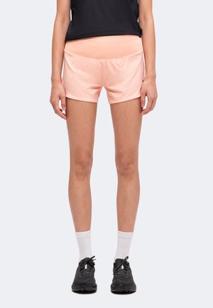 Femme portant un short de sport rose clair, un haut noir, des chaussettes blanches et des chaussures de course noires, debout devant un fond uni.