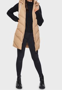 Beige Steppweste mit Kapuze, gestepptem Design und Frontreißverschluss. Trägt man über einem dunklen Grobstrickpullover und schwarzen Skinny-Jeans, kombiniert mit schwarzen Stiefeln.