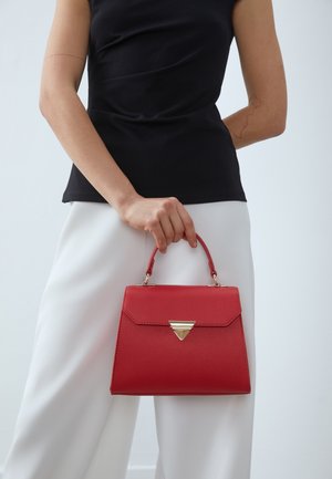 Rød struktureret håndtaske med en trekantet guldaccent, der har en top-håndtag og tekstureret overflade, båret af en person i hvide bukser.
