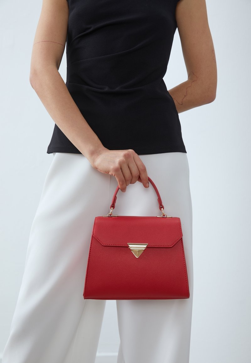 Czerwona torebka strukturalna z trójkątnym złotym akcentem, z uchwytem u góry i teksturowaną powierzchnią, trzymana przez osobę w białych spodniach.