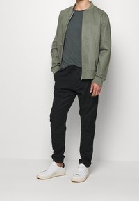 Olivfarbener Bomberjacke, dunkelgraues T-Shirt, schwarze schmal geschnittene Hose und weiße Sneakers. Einfaches Design mit elastischen Bündchen und Taille; Stoffstruktur sichtbar.
