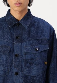 Mörkblå denimskjorta med ett strukturerat mönster, utrustad med två bröstfickor och svarta knappar. Standardkrage och synliga söm detaljer.