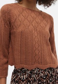 Brązowy sweter z dzianiny z diamentowymi i koronkowymi wzorami, noszony na spódnicy w print w panterkę, odsłaniający środek torsu i częściowo ramiona.