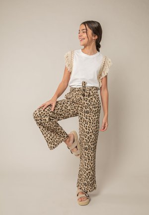 Jeune fille aux cheveux tressés portant un haut blanc à franges et un pantalon large imprimé léopard, debout avec une jambe levée, souriant sur le côté.