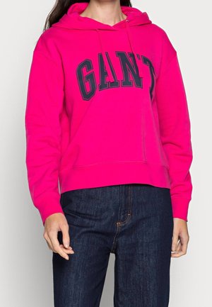 Felpa rosa con il logo "GANT" nero. Presenta un cappuccio con cordino, polsini a coste e una vestibilità comoda. Abbinata a jeans blu scuro.