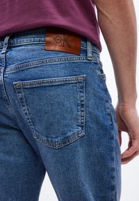 Κοντινή λήψη ατόμου που φοράει μπλε τζιν παντελόνι με καφέ δερμάτινο ταμπελάκι Calvin Klein και μωβ πουκάμισο.