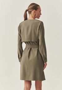 Olivengrünes Kleid mit langen Ärmeln, tailliertem Gürtel und kurzgeschnittenem, blusentypischem Oberteil. Aus glattem Stoff mit einem schlichten Design gefertigt.