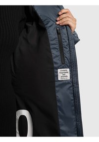 Unutrašnjost tamnoplave jakne, glatke teksture, sadrži crnu podstavu s džepom na patentni zatvarač. Oznaka glasi: "Topli zagrljaj od tkanine."