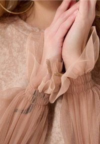 Mains jointes près du visage, portant des manches en mesh beige transparent et volanté sur un haut beige texturé avec des boucles d’oreilles dorées visibles.