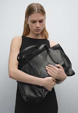 Borsa tote in pelle nera con manici allungati, texture liscia e design spazioso, tenuta vicino al corpo su uno sfondo neutro.