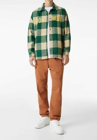 Grün-beige karierte Flanellhemd mit "NEW YORK" Schriftzug, kombiniert mit orangefarbener Cargo-Hose und weißen Sneakers. Lässiges Outfit-Design.