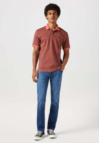 Czerwona koszulka polo z kontrastowym pomarańczowym kołnierzykiem i rękawami, zestawiona z niebieskimi dżinsami i czarnymi sneakersami. Model stoi wyprostowany z rękami w kieszeniach.