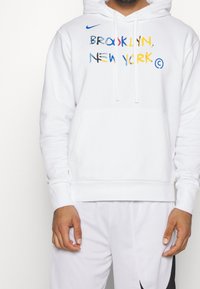 Felpa bianca Nike con cappuccio, caratterizzata da testo multicolore "BROOKLYN, NEW YORK", tasca a canguro frontale e cappuccio regolabile con cordino.