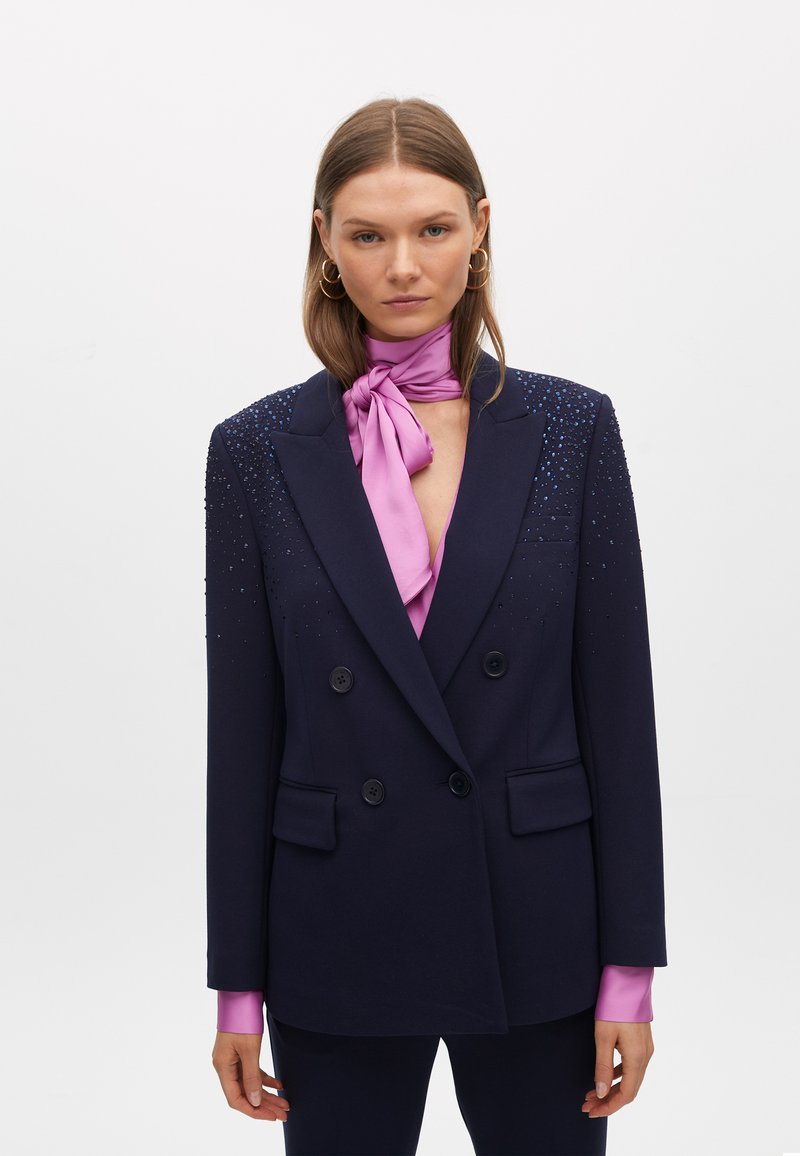 Blazer doppiopetto blu navy con decorazioni sulle spalle, dotato di sciarpa al collo in satin rosa e maniche lunghe con polsini rosa.