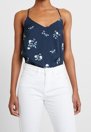 Marineblaues ärmelloses Top mit floralem Muster und dünnen Trägern, kombiniert mit hoch taillierten weißen Jeans an einer Person mit braunem Hautton.