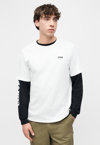 Νεαρός με σγουρά καστανά μαλλιά, φοράει στρωμέν α μαύρα μακρυμάνικα κάτω από ένα λευκό μπλουζάκι με το λογότυπο "CONS" και χακί παντελόνια, κοιτάζει δεξιά.