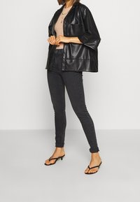 Chaqueta oversize de cuero sintético negra, top beige acanalado, pantalones ajustados oscuros y chanclas de tacón negras. Diseño simple con un bolsillo en el pecho.