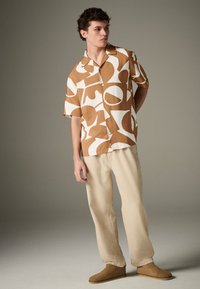 Trumpomis rankovėmis marškiniai su sagutėmis rudos ir gelsvai baltos abstrakčios raštų, derinami su šviesiai smėlio spalvos kelnėmis ir rudais zomšos bateliais be užsegimo.