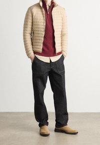 Beige vadderad jacka, burgundy stickad tröja, ljus skjorta, mörk denimbyxor och beige stövlar. Släta texturer med synliga sömmar.