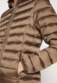 Nahaufnahme einer Person, die eine braune gepolsterte Steppjacke mit Frontreißverschluss trägt und die Hand in einer seitlichen Reißverschlusstasche hat.