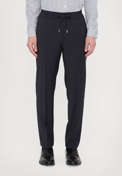 Los pantalones negros con cordón tienen un corte relajado, una tela suave y un brillo sutil. Se combinan con una camisa a rayas y zapatos negros.