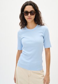 Himmelblaues, geripptes Kurzarmoberteil mit runder Ausschnitt. Glatte Textur und figurnahe Passform. Kombiniert mit einer cremefarbenen Hose. Schildpatt-Sonnenbrille.