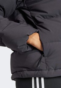 Kurtka puchowa o matowej czarnej powierzchni, z fakturowanym wykończeniem i elastycznym sznurkiem ściągającym na dole. Ręka częściowo w kieszeni.