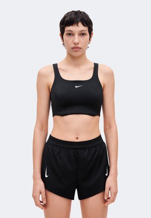 Un mannequin aux cheveux courts se tient face à l’objectif, vêtu d’une brassière de sport Nike noire et d’un short assorti, sur un fond clair uni.