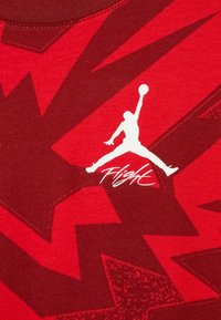 Rode T-shirt met abstracte donkerrode patronen. Beschikt over een witte silhouetlogo en het woord "Flight" in cursief. Katoenen materiaal, klassieke ronde hals ontwerp.