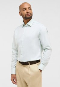 Helle mintgrüne langärmelige Bluse mit einem Button-Down-Kragen und subtiler Textur, kombiniert mit beigen Hosen und einem braunen Gürtel.