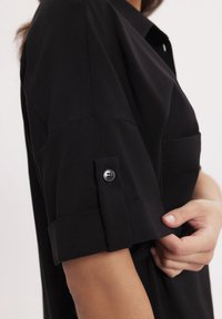 Czarna koszula z krótkim, podwiniętym rękawem i zapięciem na guzik. Materiał wydaje się gładki, o prostym designie i bez widocznych wzorów.