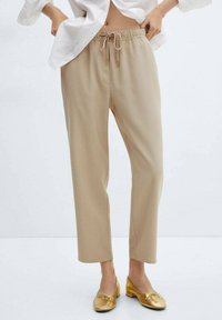 Pantalones jogger beige con cintura elástica y cordón, con un corte relajado y piernas rectas, combinados con zapatos planos dorados.