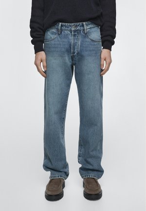 Person trägt hellblaue Jeans mit geradem Bein, braune Loafers und einen schwarzen Langarm-Pullover und steht vor einem schlichten hellen Hintergrund.