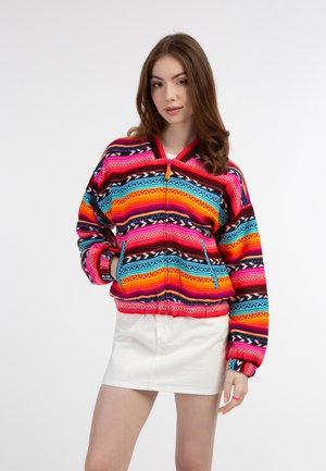 Kleurrijke ritjack met gedurfde veelkleurige patronen in roze, blauw en oranje; heeft een kraag en elastische manchetten. Witte rok eronder.