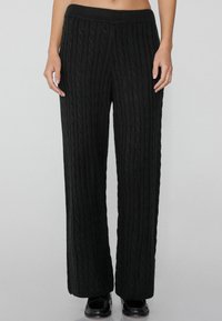 Zwarte kabelgebreide wijde broek met een geribbelde elastische tailleband. De stof heeft een gestructureerd patroon, wat zorgt voor een comfortabele en losse pasvorm.