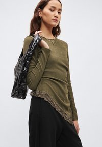 Olive žalia, siaurėjanti ilgomis rankovėmis palaidinė su apvalia apačia, puošta nėrinių kraštu apačioje, derinama su juoda lakuotos odos striuke.