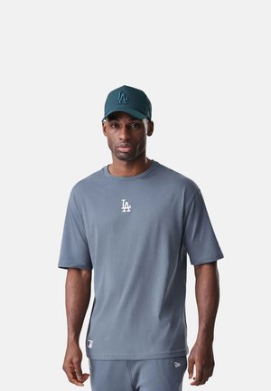 Uomo che indossa un cappellino, una maglietta e pantaloni grigi coordinati con il logo LA, in piedi davanti a uno sfondo grigio chiaro uniforme.