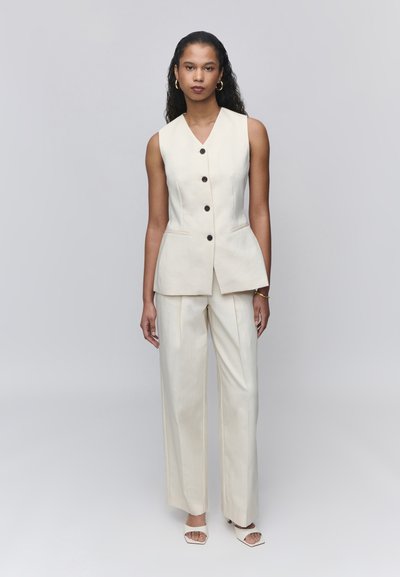 Femme aux longs cheveux bouclés porte un gilet sans manches crème boutonné, un pantalon large assorti, des boucles d’oreilles créoles dorées et des talons blancs ouverts à l'avant.