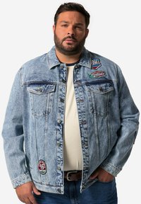 Chaqueta de mezclilla de color azul claro con dos bolsillos en el pecho, varios parches coloridos y cierres de botón. Usada sobre una camisa de color claro.