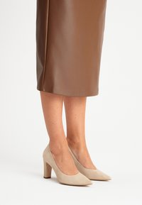 Pumps beige in suede con tacco alto, punta affilata, tacco chunky e texture liscia, abbinati a una gonna a tubino in pelle marrone.