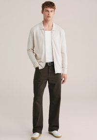 Svetlobéžový kardigán cez biele tričko, doplnený tmavozelenými nohavicami. Obuv je biela s svetlohnedými podrážkami.