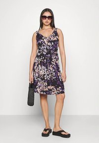 Vestido floral en negro, púrpura y durazno; sin mangas, corpiño ajustado, falda fluida. Combinado con sandalias de plataforma negras y un bolso tejido.
