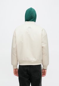 Kreemivärvi pommitaja jope rohelise kapuutsiga, soonikkoes varrukate ja äärega. Seljal on väike logomärgiga silt. Kantud tumedate teksade peal.