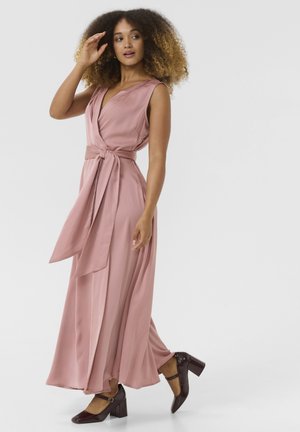 Maxi dress in soft pink fabric with a wrap design and self-tie belt. Features sleeveless cut and flowing silhouette. Worn with dark heeled shoes.