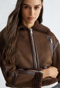 Giacca marrone con un colletto morbido e marrone e dettagli laterali. Presenta zip argentate e rifiniture in pelle. Finitura morbida e testurizzata.