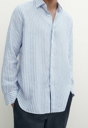 Man met een lichtblauw-wit verticaal gestreept overhemd met lange mouwen, handen in de zakken van een donkere broek.