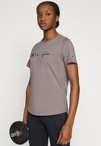 Gray cotton t-shirt with short sleeves, featuring embroidered "Champion" logo in black. Smooth texture, casual fit, paired with a black dumbbell.