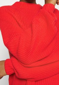 Rode gebreide trui met een geribbelde textuur, voorzien van een hoge halslijn en lange, volumineuze mouwen. Strak aansluitende manchetten accentueren de mouwen.