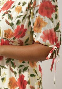 Kwiatowa sukienka w czerwone, pomarańczowe i zielone wzory na kremowym tle. Krótkie bufiaste rękawy związywane na mankietach.